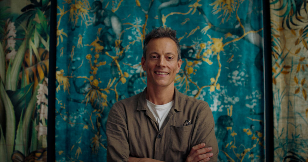 Adam Ellis smiling into camera in front of a hand-painted wallpaper, from KYN's micro-documentary series