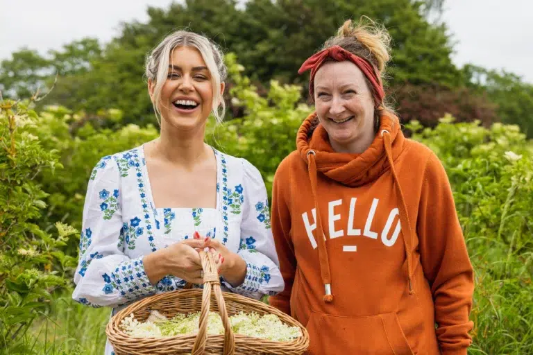 An image of Daisy Payne holding a basket of Elderflower flowers on Belvoir Farm standing next to a farm worker.