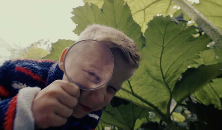 An image of a young boy wearing a striped fleece looking at the camera through a magnifying glass.