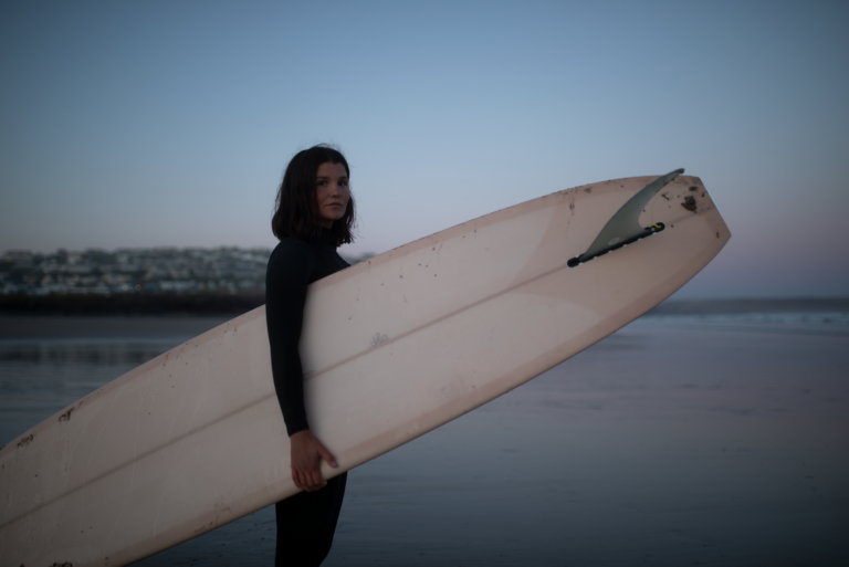 An image of illustrator and surfer Clara Jonas in Cornwall holding a surf board at golden hour on Polzeath beach.