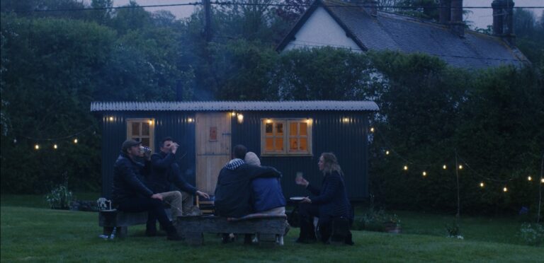 An image of a plank bridge hut with fairy lights all of it. There is a group of people having a bonfire infront of it.