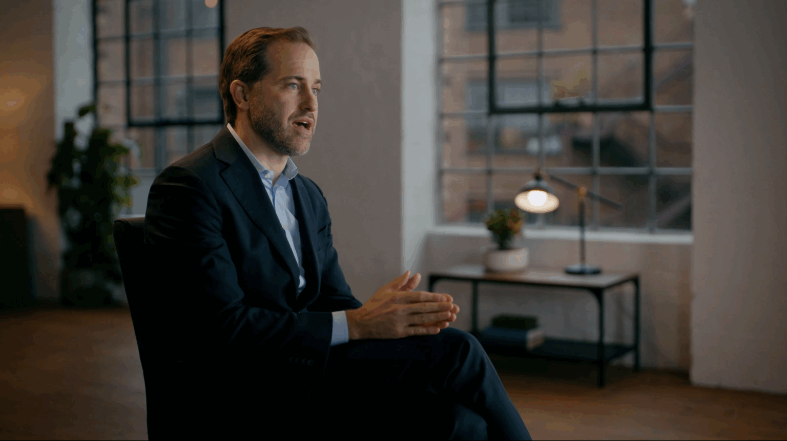 Hire a DOP in London 6 A man in a suit mid-answer in a lit interview, a lamp glowing by the window behind him