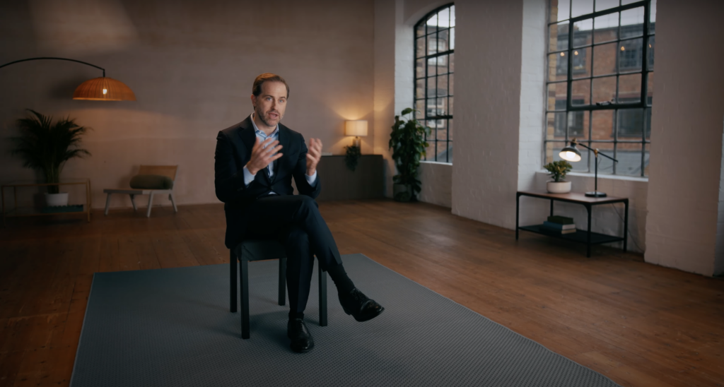 A wide interview frame in a loft studio: one chair on a grey rug, warm practical lamps and tall arched windows