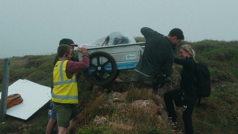 A behind the scenes shot from our recent cohere shoot. we see a film crew in Cornwall lifting a trolly of film kit over a dry stone wall to get the equipment onto set.