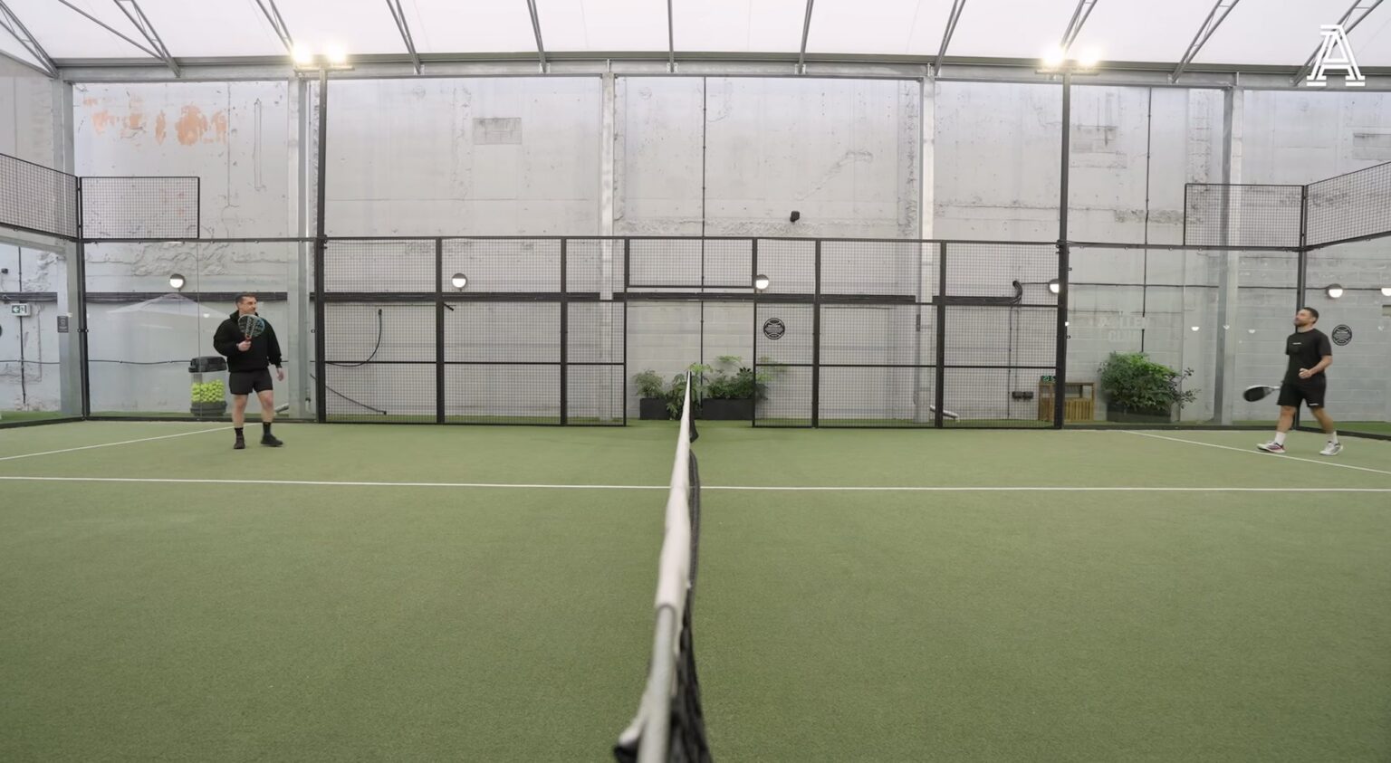 A low wide shot down the net of a covered padel court, Gary Neville at one end and David Ornstein at the other